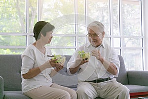 Smiling Asian senior man and woman sitting on sofa drinking glasses of milk. Mature couple enjoying healthy salad food while