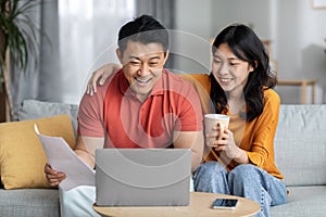 Smiling asian spouses paying bills online, using laptop