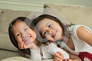 Smiling asian toddler sisters laying on stomach on couch