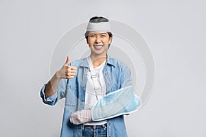 Smiling asian woman with broken head and wearing soft cast due to broken arm while showing thumbs up isolated on white background