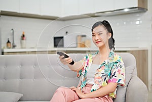 Smiling asian woman on couch changing tv channel at home in the living room