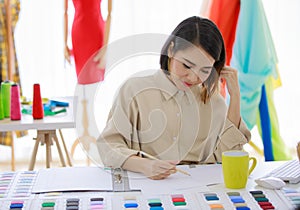 Smiling Asian woman dressmaker is sitting  sketch dress that she has to cut for the customer in the Tailor shop Concept Fashion