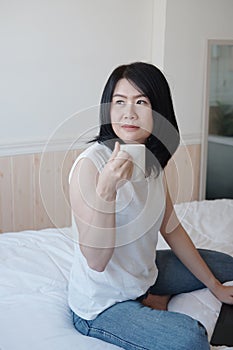 Smiling Asian woman is drinking a cup of coffee and working with laptop in bedroom on holiday. She is online working from home