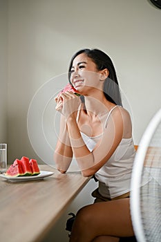 A smiling Asian woman enjoys her fresh watermelons while sitting in front of a fan