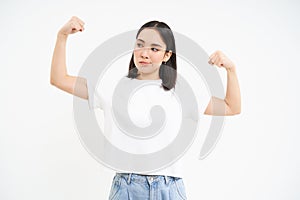 Smiling asian woman, flexing biceps, looking sassy and strong, showing her strength muscles, white studio background
