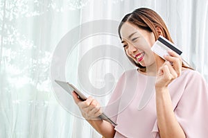 Smiling asian woman holding discount credit card in hand paying for shopping online at tablet in home, Young casual lifestyle