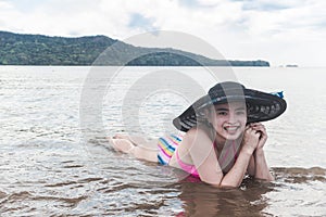 A smiling asian woman lying in the sand and surf in shallow waters at the beach. Wearing a large sun hat