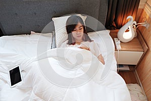 Smiling Asian woman is relaxing and laying and online internet with smartphone before sleep and good night in bedroom on holiday.