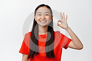 Smiling asian woman showing okay, ok sign and looking satisfied, recommending good quality, standing against white