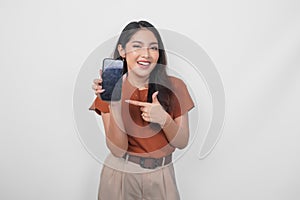 Smiling Asian woman showing and pointing to the copy space on her smartphone wearing brown shirt  by a white background