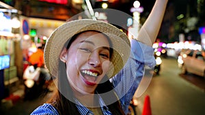 Smiling Asian woman in straw hat greeting with a wave, city nightlife sparkling in the background
