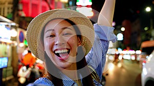 Smiling Asian woman in straw hat greeting with a wave, city nightlife sparkling in the background