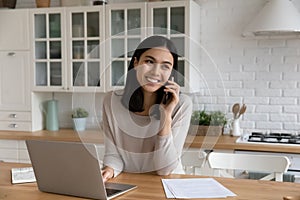 Smiling Asian woman talking on phone, using laptop in kitchen