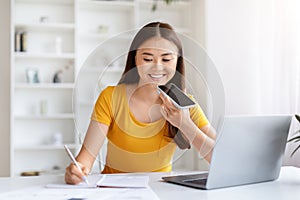 Smiling asian woman using smartphone for voice command while writing notes