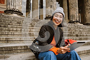 Smiling asian woman using tablet computer while sitting on stairs at old city street