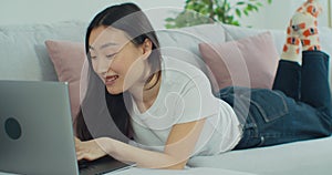 Smiling Asian woman working from home lying on the comfortable sofa. Woman using laptop notebook looking at screen