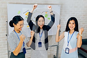 Smiling Asian young and mature business women standing in line with arms raised up gesture in meeting room with excited