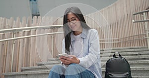 Smiling Asian young student female seats on the stairs and using smartphone chating and typing message