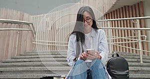 Smiling Asian young student female seats on the stairs and using smartphone chating and typing message