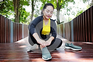 Smiling Asian young woman in black sportwear resting and tying Shoelace on wooden bridge before exercise and running in garden
