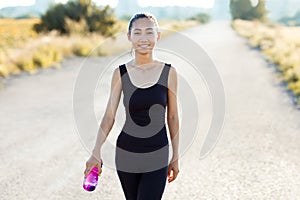 Smiling athletic woman walking on rural road and carrying water