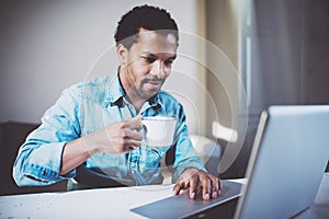 Smiling attractive African man making video conversation via laptop with partners while drinking white cup black tea in