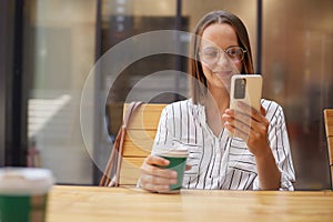 Smiling attractive brown haired businesswoman in formal attire while using smartphones drinking coffee in cafe reading emails