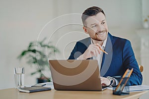 Smiling attractive businessman sitting in front of laptop with pencil in hand and looking aside
