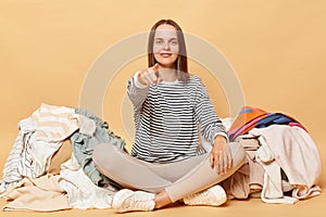 Smiling attractive Caucasian woman posing near heap of multicolored unsorted clothes isolated over beige background pointing to