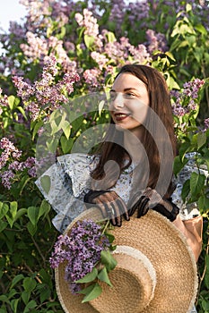 Smiling attractive caucasian woman in retro clothes holding a boatman& x27;s straw hat in a blooming lilac garden.
