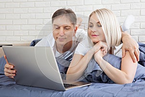 Smiling attractive couple lying on bed using laptop communicating online at home, happy man and woman typing on computer