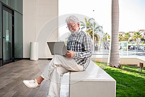 Smiling attractive elderly businessman holding laptop computer on his knees sitting out of his office. Mature attractive man