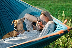 Smiling attractive European man in sunglasses is resting in hammock with his cute little dog