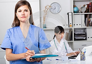 Smiling attractive female doctor writing notes on clipboard in office with colleague