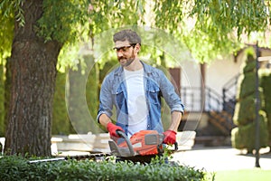 Smiling attractive male worker trimming shrub with hedge cutter in park.