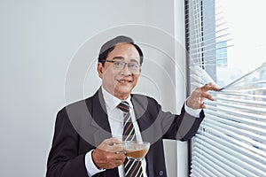 Smiling attractive mature man wearing glasses leaning on the sill of a large bright window with copy space looking at the camera