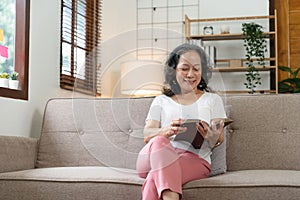 smiling attractive retired woman reading book while sitting on sofa at home.