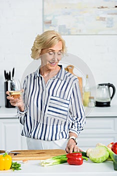 smiling attractive senior woman holding glass of white wine in kitchen and looking