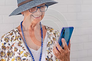 Smiling attractive senior woman wearing blue hat using mobile phone - standing against a white brick wall - elderly people