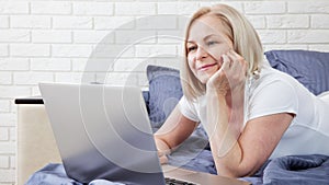 Smiling attractive woman lying on bed using laptop communicating online at home, happy girl typing on computer, enjoying