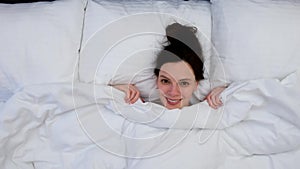 Smiling attractive young brunette woman hiding under the blanket laying in bed.