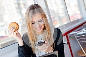 Smiling attractive young business woman eating a delicious burger in a cafe or restaurant holding the mobile cell phone chatting