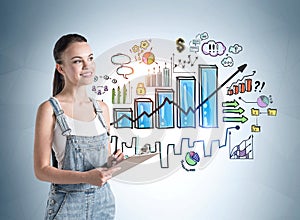 Smiling attractive young woman in denim overalls holding a planner and standing near concrete wall with creative colorful bar