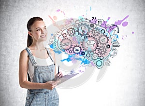 Smiling attractive young woman in denim overalls holding a planner and standing near concrete wall with creative colorful idea