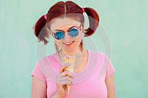 Smiling audacious young woman with pigtails hairstyle holds ice-cream in waffle cone, it reflects in her sunglasses. Urban style