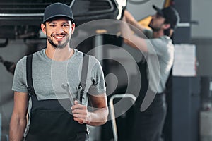 smiling auto mechanic holding wrenches while colleague working