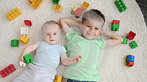 Smiling baby boy with older brother lying on carpet next to heap of colroful toys and looking up in camera. Concept of
