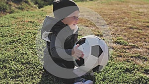 Smiling baby boy with a soccer ball at football field. Portrait of a little child sitting at stadium with a ball. Future