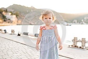 Smiling baby girl 1-2 year old wear striped dress and holding walk over wooden pier at sea shore outdoors. Summer season.