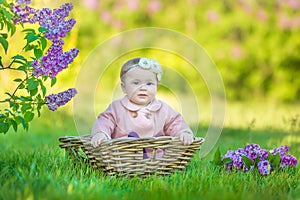 Smiling baby girl 1-2 year old wearing flower wreath, holding bouquet of lilac outdoors. Looking at camera. Summer spring time.